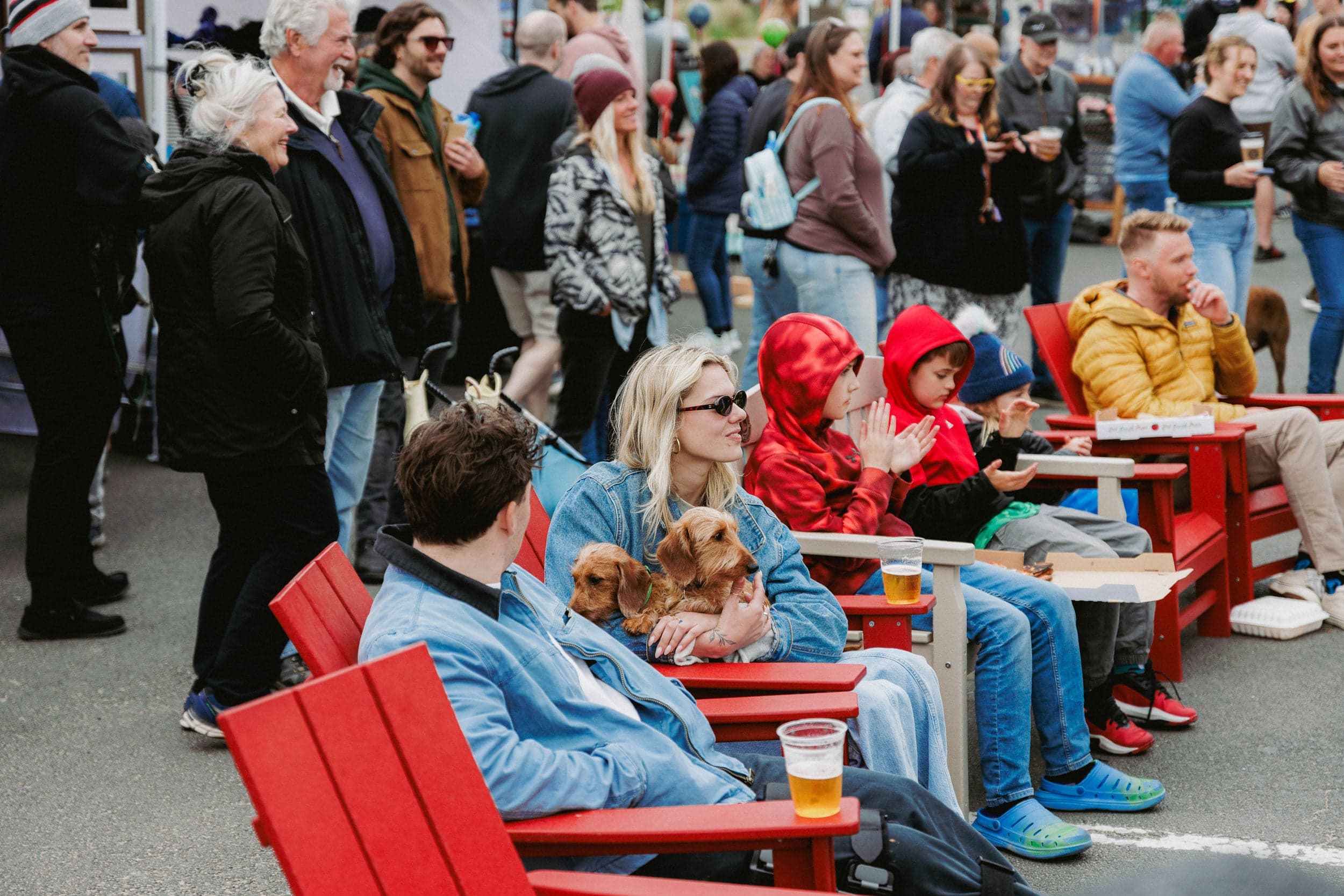 A crowd enjoys an outdoor event. People sit on red chairs, some holding drinks, while a woman cuddles two dogs. The scene feels lively and social.