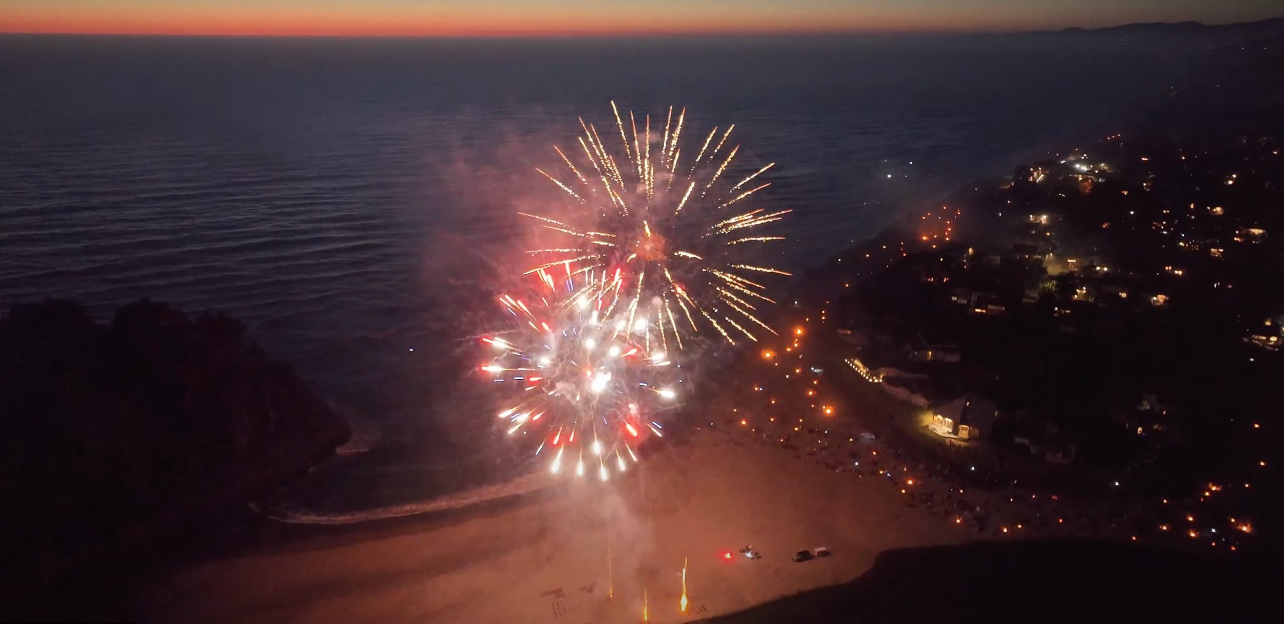 Aerial view of colorful fireworks bursting over a beach at dusk. Distant city lights twinkle on the right, creating a festive and celebratory atmosphere.