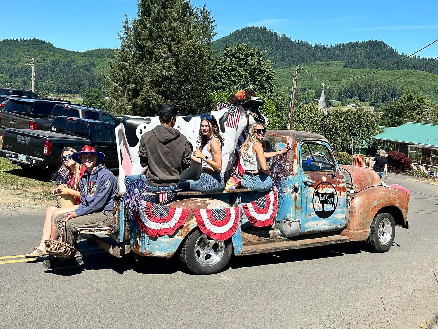 A vintage truck adorned with American flags and bunting carries smiling people in a parade. A large cow model is on the truck, set against a scenic, green hilly backdrop.