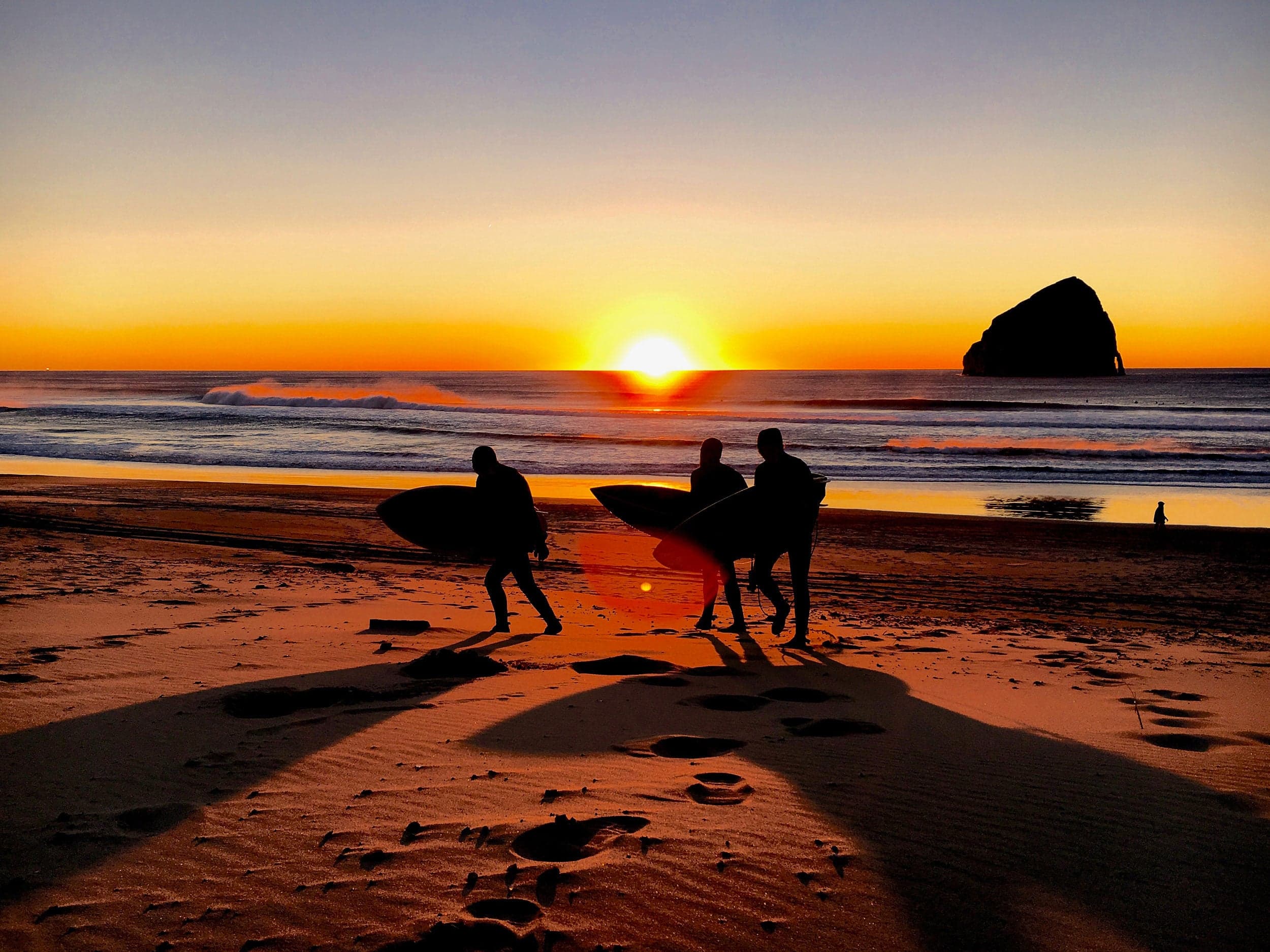 Silhouetted surfers carrying boards across a sandy beach at sunset, with a glowing sun on the horizon, gentle waves, and a distant rock formation.