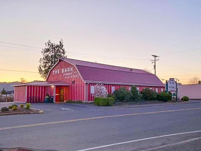 Red barn-style building with white trim, labeled "The Barn," at sunset. Surrounded by shrubs, it sits next to a road with a small sign nearby.