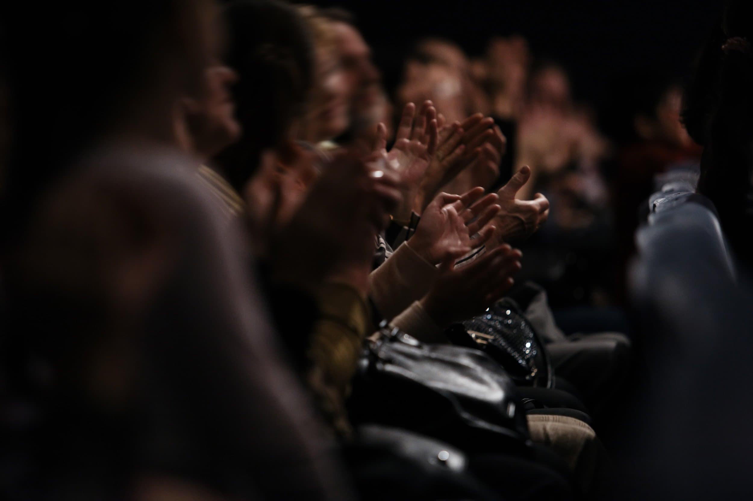 Audience members are clapping in a dimly lit theater, conveying appreciation and excitement. The focus is on their hands, with a blurred background.